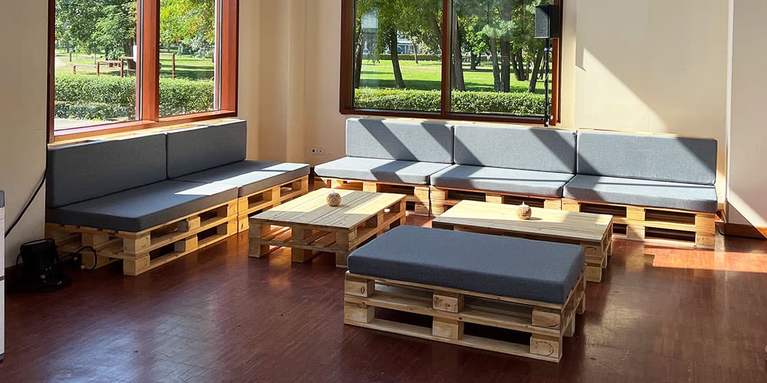 Lounge-Bereich aus Palettenmöbeln in einem Tagungsraum – mehrere Sitzbänke mit grauen Kissen und zwei Loungetische bei Tageslicht