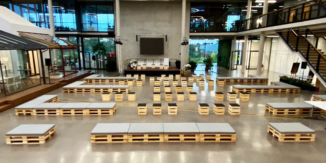 Großveranstaltung in Industrie-Atrium mit zahlreichen Palettenbänken und Hockern als Bühnenbestuhlung – Konferenz-Setup im Palettenstil
