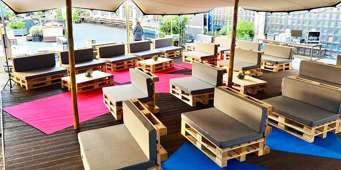 Outdoor-Lounge unter Pavillon mit Palettenbänken, grauen Sitzkissen und farbigen Teppichen – Festival-Sitzgruppen im Freien
