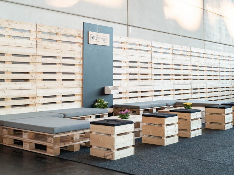 Palettenwand mieten Berlin – Sitzmoment-Loungebereich auf Messe mit Palettenliegen, grauen Sitzauflagen, Hocker-Cubes und großer Palettenwand-Rückwand mit Sitzmoment-Logo