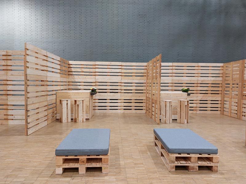Palettenwand mieten Leipzig – Kompletter Eventraum mit umlaufender Palettenwand, zwei Stehtischen mit Blumen und zwei Palettenliegen mit grauen Polstern