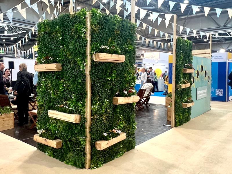 Pflanzenwand mieten Berlin – kabinenartiger Pflanzenwand-Würfel mit grüner Begrünung und Holz-Pflanzkästen auf dem 'We are Europe'-Messestand mit blauen Wimpelketten