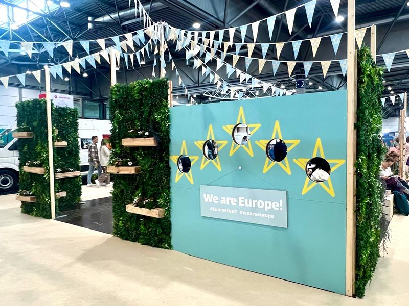 Pflanzenwand mieten Hamburg – grüne Pflanzenwand mit Holz-Pflanzregalen rahmt 'We are Europe'-Messestand mit gelben Sternen ein, Wimpelketten in Pastellblau