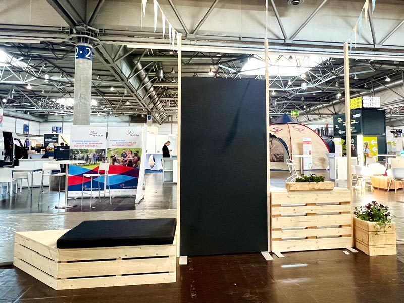 Tafelwand mieten Berlin – große freistehende Kreidetafel-Wand auf Messe, flankiert von Palettenliege mit schwarzer Auflage und Holz-Pflanzkästen mit Blumen